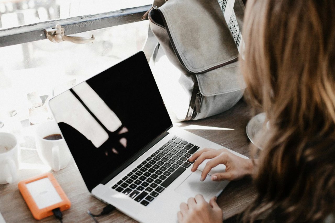 A young woman works remotely at a café, using her laptop and external hard drive.